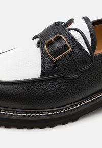 Mocassin en cuir noir et blanc avec un dessus texturé, proposant une boucle en métal et des coutures contrastées le long de la semelle, avec une semelle en caoutchouc.