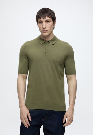 Ung mand med kort mørkt hår iført en tætsiddende olivengrøn kortærmet polo og mørkeblå jeans, stående mod en ensfarvet baggrund.