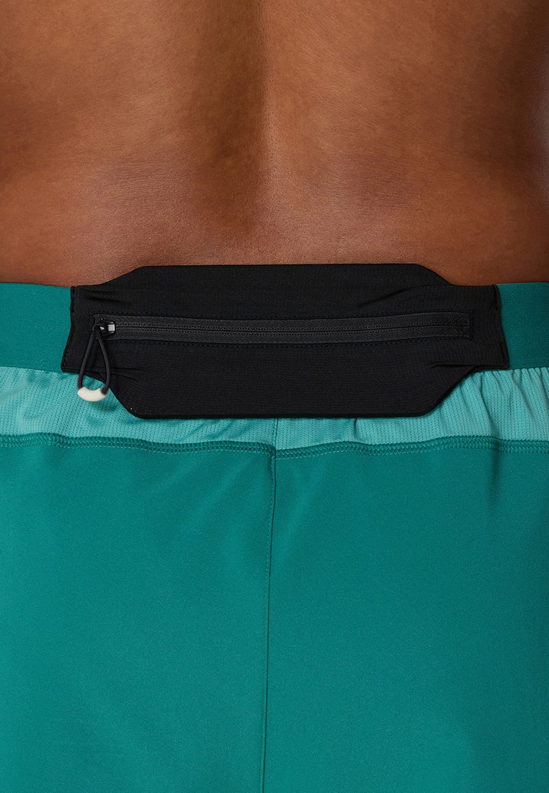 Zwarte ritszak bevestigd aan de tailleband van groene shorts, gemaakt van gladde stof met een strakke pasvorm en minimale naaddetails.