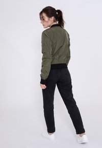 Giacca bomber verde con maniche e orlo a coste neri, caratterizzata da una trama liscia. Combinata con jeans neri e sneakers bianche. Design minimalista.