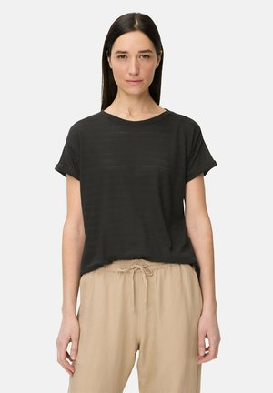 Kvinde iført en sort kortærmet tekstureret skjorte, stoppet ned i beige bukser med snoretræk, stående mod en ensfarvet lys baggrund.