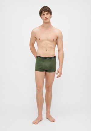 Πράσινες βαμβακερές μποξερίτσες με μαύρη ζώνη λογότυπου Moschino, στενή σχεδίαση, λεία υφή και μήκος μέχρι το μέσο του μηρού σε όρθιο άνδρα μοντέλο.