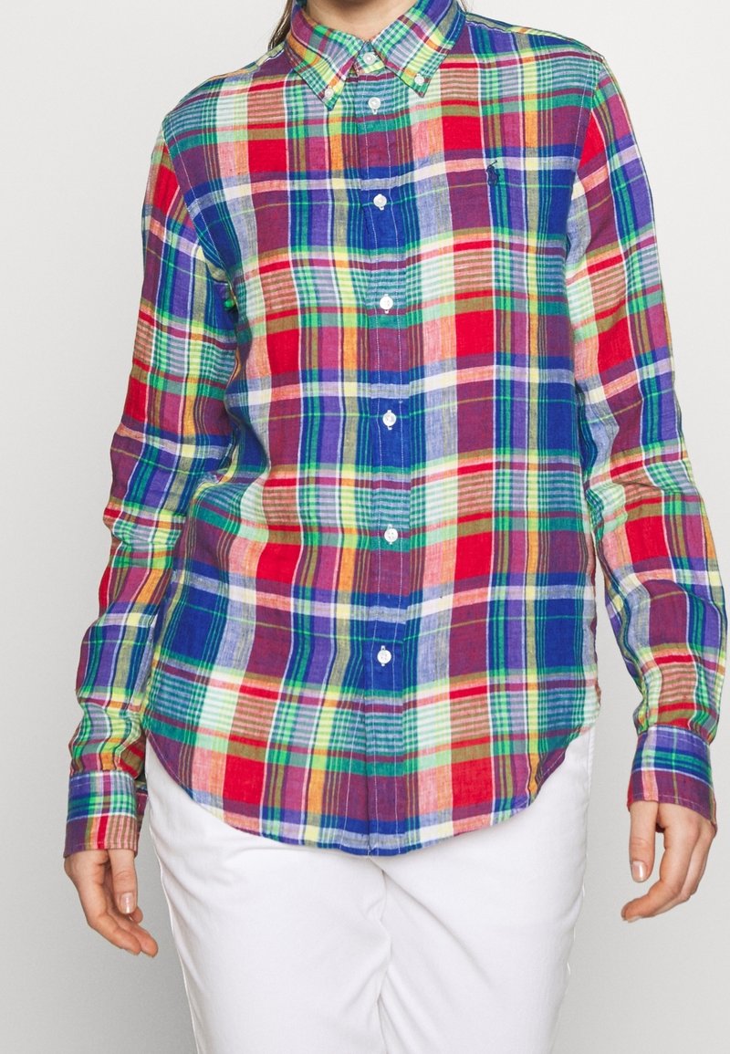 Personne portant une chemise colorée à carreaux à manches longues et un pantalon blanc, cadrée au niveau du cou et de la taille.