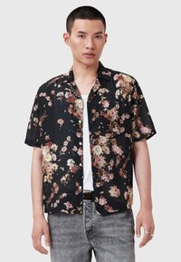 Bărbat tânăr îmbrăcat cu o cămașă neagră cu mânecă scurtă, cu imprimeu floral în nuanțe de roz și bej, tricou alb pe dedesubt, blugi gri deschis și inele argintii pe degete.