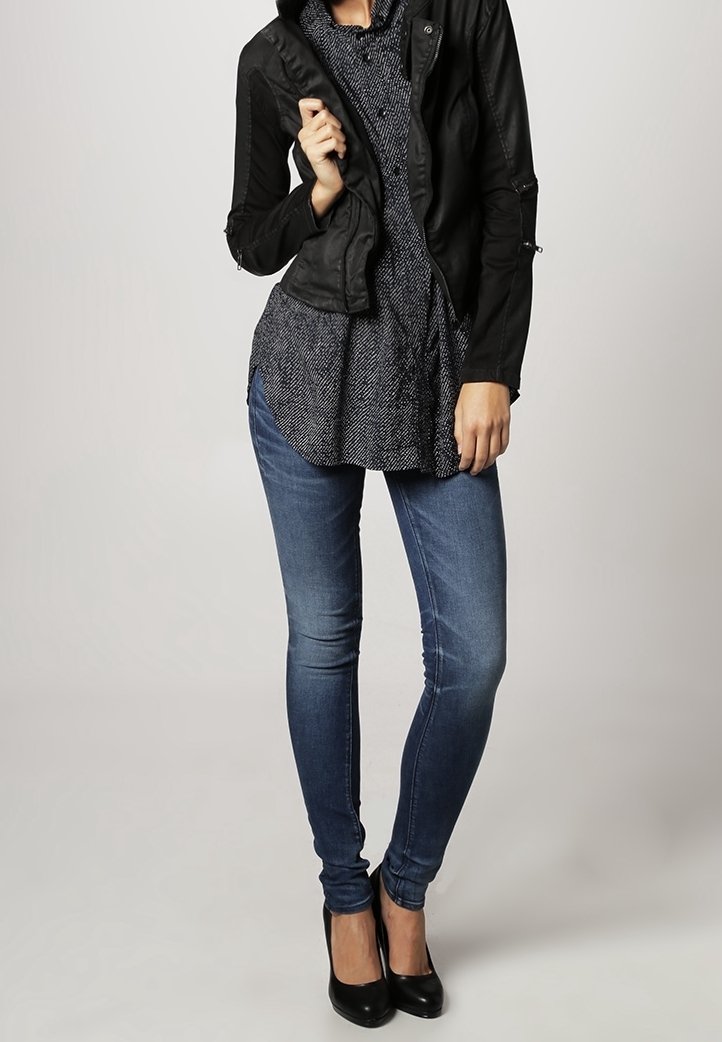 Veste noire courte sur une chemise à motifs bleu marine, associée à un jean bleu ajusté et des talons noirs. Textures lisses et coupe ajustée partout.