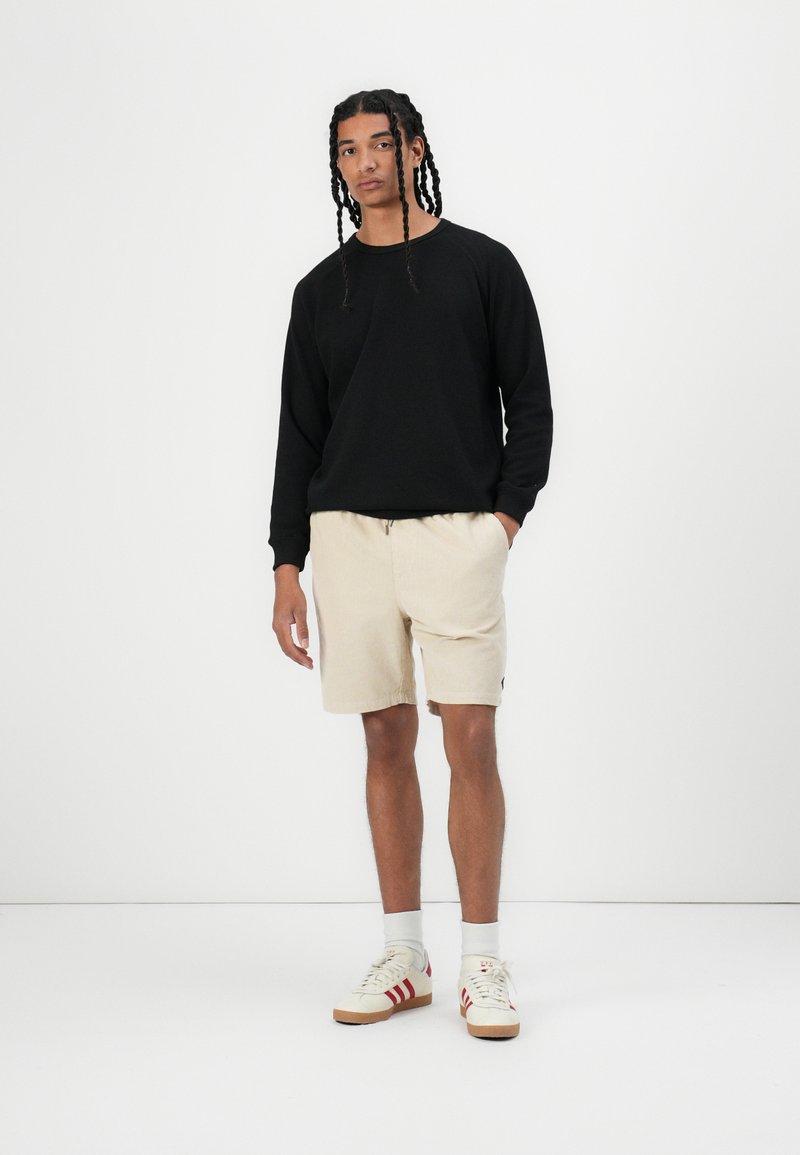 Sweatshirt noir à manches longues, shorts en velours côtelé beige et baskets beige avec des accents rouges. Design simple, coupe décontractée.