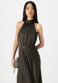 Tamsiai alyvuogių spalvos halter-aiste kurta su susirinkusia juosmeniu, pagaminta iš tekstūrinio audinio. Modelis laiko metalinę, dekoruotą karoliukais rankinę su grandinėle.