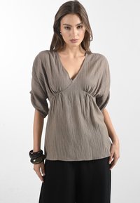 Blusa marrone chiaro con scollo a V, maniche a sbuffo e dettagli arricciati in vita; tessuto strutturato, abbinata a pantaloni neri e a una serie di braccialetti.