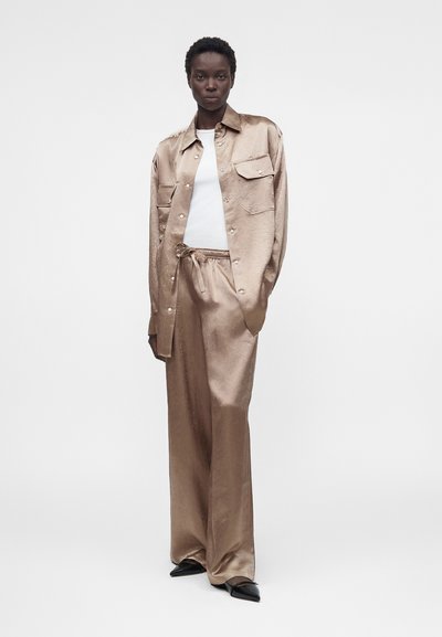 Ψηλό άτομο φορώντας άνετο μπεζ σατέν πουκάμισο και παντελόνι πάνω από λευκό τοπ με μαύρα μυτερά παπούτσια, στέκεται μπροστά από απλό φόντο.