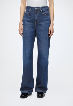 Person iført blå jeans med høj talje, lige ben og klassisk fem-lomme design samt sorte sko med firkantet tå, stående mod en hvid baggrund.
