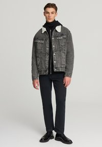 Giovane uomo in piedi con indosso una giacca di jeans grigia foderata in sherpa, un dolcevita nero, pantaloni scuri e scarpe in pelle nere su uno sfondo semplice.