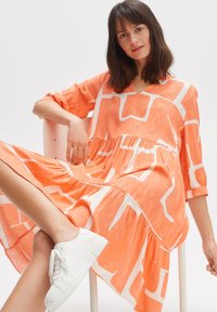 Orangekleid mit weißem geometrischem Muster, leichtem Stoff, V-Ausschnitt, drei Viertelärmeln und schichtetem Rock. Kombiniert mit weißen Sneakers.