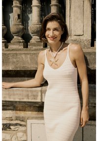 Abito bianco lavorato a maglia con spalline sottili, caratterizzato da un motivo testurizzato. Accentuato da una collana a forma di stella marina. Sullo sfondo ci sono balaustre in pietra.
