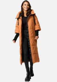 Lange, puffy oranje jas met zwarte accenten, voorzien van een capuchon en ritssluiting, gecombineerd met zwarte leggings en stevige zwarte laarzen.