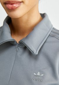Grå zip-up jakke med teksturert stoff, med krage og Adidas-logoen i lys broderi på venstre side.