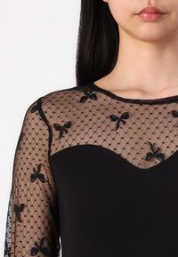 Schwarzes Langarmshirt mit einer transparenten, gepunkteten Mesh-Oberlage und schwarzen Schleifen. Der untere Teil besteht aus einem einfarbigen schwarzen Stoff mit einem gewellten Ausschnitt.