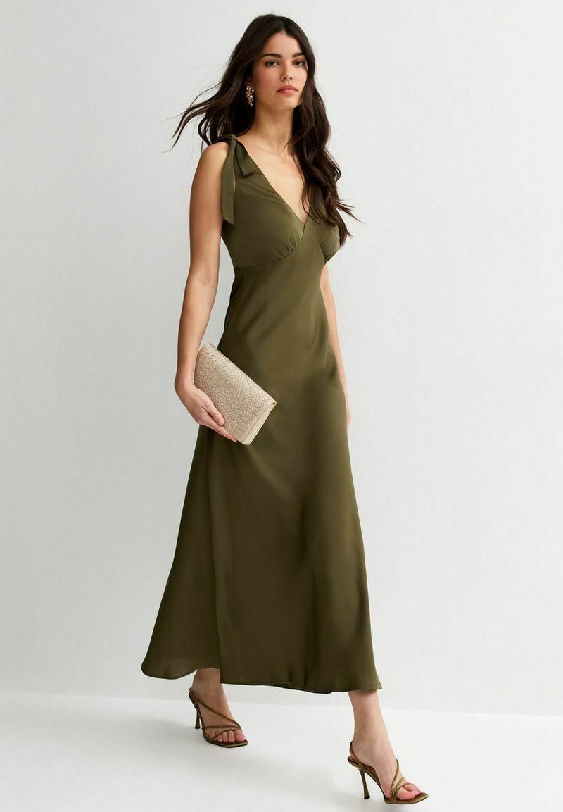 Vrouw die loopt en een olijfgroene mouwloze jurk met schouderstrik draagt, een beige clutch vasthoudt en bruine hoge hakken sandalen draagt.