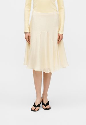 Γυναίκα με κρεμ midi φούστα με φουσκωτή γραμμή, ταιριαστό μακρυμάνικο τοπ, ασημένια βραχιόλια και μαύρα ψηλοτάκουνα σανδάλια με ανοιχτά δάχτυλα, στέκεται σε λευκό φόντο.