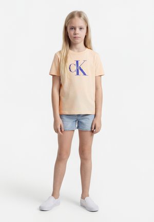 Mladé dievča stojí, oblečené v broskyňovom tričku Calvin Klein, svetlých riflových kraťasoch a bielych šmykľavých topánkach, na jednoduchom bielom pozadí.