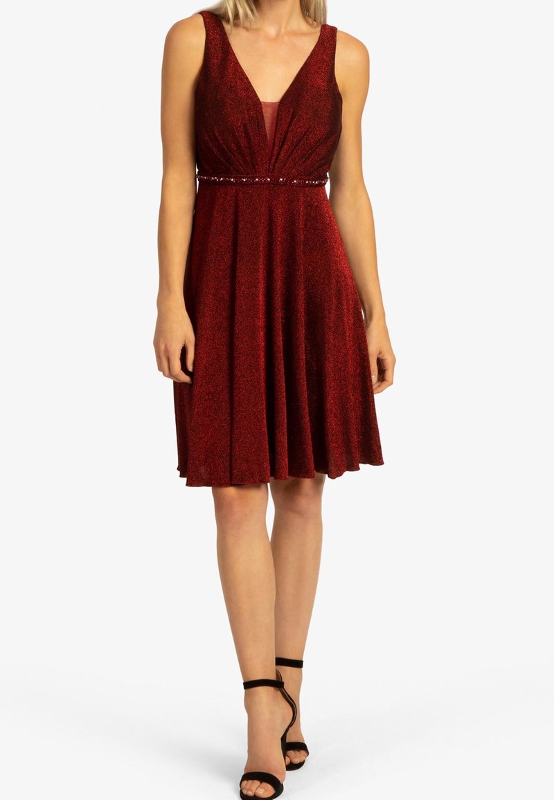 Femme portant une robe sans manches, longueur genou, rouge profond pailletée avec décolleté en V et détail de taille perlé, associée à des talons noirs à bride à la cheville.