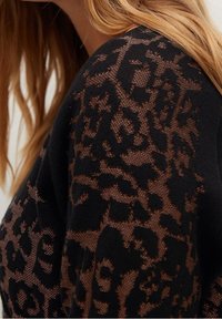 Czarny dzianinowy top z przezroczystymi brązowymi rękawami we wzór w panterkę, z misternymi wzorami i miękką teksturą, podkreślający warstwowe elementy projektu.