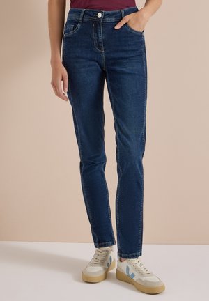 Personne portant un jean slim bleu foncé et des baskets blanches avec des touches bleues, debout devant un fond beige uni.