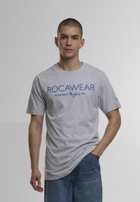 Grå bomulds-t-shirt med blå "ROCAWEAR"-tekst og "ESTABLISHED New York 1999"-logo. Korte ærmer og afslappet pasform, parret med denimshorts.