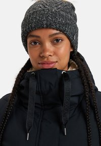 Chaqueta acolchada negra con un alto cuello y gorro de punto gris. Presenta cabello trenzado y cordones negros con puntas plateadas. Fondo claro.