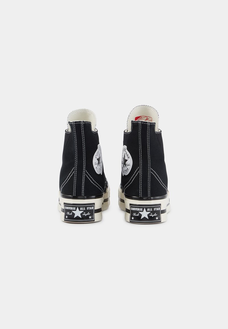 Des baskets montantes noires en toile, avec des coutures blanches, un patch logo sur le côté, et une semelle en caoutchouc avec des détails de marque.