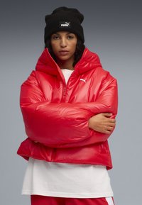 Rode pufferjack met een glanzende afwerking, voorzien van een hoge kraag en een vooraanstaande rits. Draagt over een wit shirt. Zwart gebreid beanie op het hoofd.