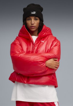 Rode pufferjack met een glanzende afwerking, voorzien van een hoge kraag en een vooraanstaande rits. Draagt over een wit shirt. Zwart gebreid beanie op het hoofd.
