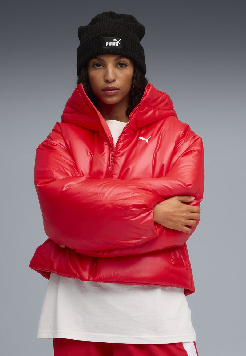 Rode pufferjack met een glanzende afwerking, voorzien van een hoge kraag en een vooraanstaande rits. Draagt over een wit shirt. Zwart gebreid beanie op het hoofd.