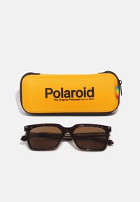 Ochelari de soare Polaroid cu lentile maronii închis, într-un cadru pătrat, asociați cu un toc rigid galben cu logo negru și închidere cu fermoar.