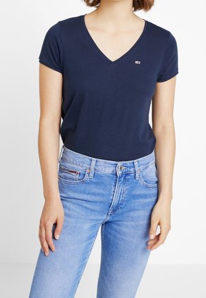 Kvinde iført en marineblå T-shirt med V-hals og korte ærmer, stoppet ned i lyseblå højtaljede jeans med forlommer og bæltestropper.