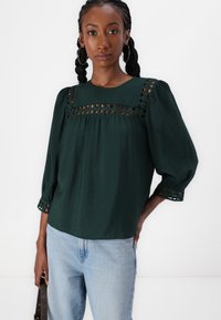 Blusa verde scuro in tessuto testurizzato, con un design a taglio sul petto e sulle maniche, abbinata a jeans blu chiaro.