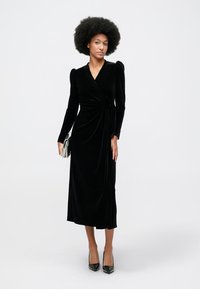 Ženska z afro frizuro, oblečena v dolgo črno žametno obleko z naborki, drži srebrno torbico in stoji ob belem ozadju.