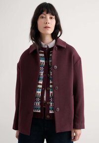Burgundowa wełniana kurtka z kołnierzem i guzikami z przodu, nałożona na sweter z wzorem z niebieskimi, zielonymi i kremowymi detalami.