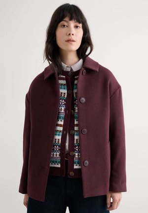 Bordeaux wollen jas met een kraag en voor knopen, gelaagd over een gebreide trui met een patroon in blauw, groen en crèmekleurige details.