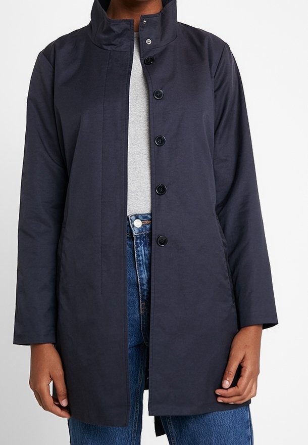 Cappotto blu navy, di lunghezza media, con colletto alto, chiusura a bottoni e maniche lunghe; realizzato in tessuto liscio, indossato sopra un top grigio e jeans.