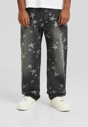 Weit geschnittene dunkelgraue Jeans mit ausgewaschenen weißen Sternmustern, kombiniert mit weißen Sneakers, sichtbar von der Mitte des Oberschenkels abwärts.