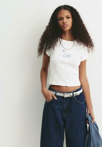 Donna con lunghi capelli ricci che indossa un crop top bianco con una grafica di cigno, jeans larghi scuri, cintura bianca, mentre tiene in mano una giacca di denim.