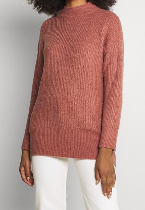 Vrouw draagt een stoffig roze geribbelde gebreide trui met lange mouwen, gecombineerd met een witte broek tegen een effen achtergrond.