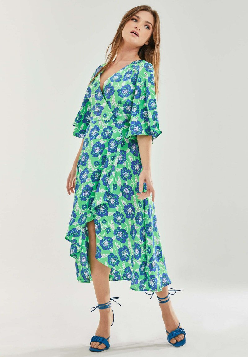 Floral wrap dress in green with blue flowers, featuring flutter sleeves, a V-neck, and a side slit. Paired with blue heeled sandals.