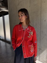 Piros kardigán fehér varrással, kígyó és nap hímzett mintákkal a zsebeken, puha kötött anyagból készült.