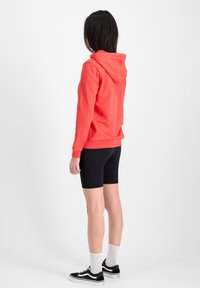 Sudadera con capucha de color coral y corte relajado, con puños acanalados, combinada con pantalones cortos de ciclismo negros. Modelo que lleva calcetines blancos y zapatillas negras.