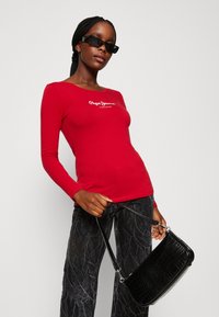 Camicia rossa a maniche lunghe con scollo rotondo e logo bianco. Borsa nera con stampa coccodrillo a rilievo, dotata di tracolla, abbinata a pantaloni scuri texturizzati.
