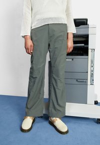 Pantalones cargo grises con bolsillos laterales, combinados con zapatos de un beige claro. Los pantalones tienen un corte holgado y un brillo sutil, llevados con un suéter de punto blanco.