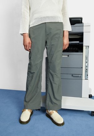 Pantalones cargo grises con bolsillos laterales, combinados con zapatos de un beige claro. Los pantalones tienen un corte holgado y un brillo sutil, llevados con un suéter de punto blanco.