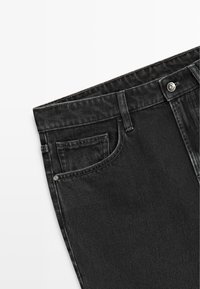 Sort denimjeans med forlomme og bæltestrop, synlig metal knap og nitte-detalje på hvid baggrund.