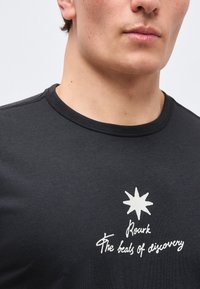 Άνδρας που φοράει μαύρο μπλουζάκι με στρογγυλή λαιμόκοψη, με ένα λευκό αστέρι και το κείμενο «Rosark The beats of discovery» στο στήθος.